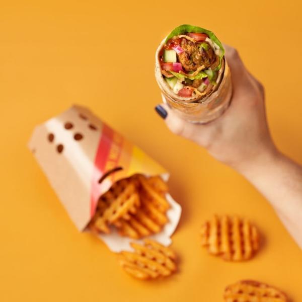 Hand holding a veggie wrap above a yellow table loaded with waffle fries
